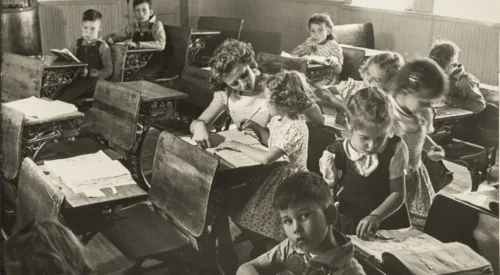 Black and white photograph of a primary school classroom in 1948. Light-skinned children sit at dark wood desks attending to books and papers or looking directly into the camera. At the center, the young teacher assists a girl with her lesson.