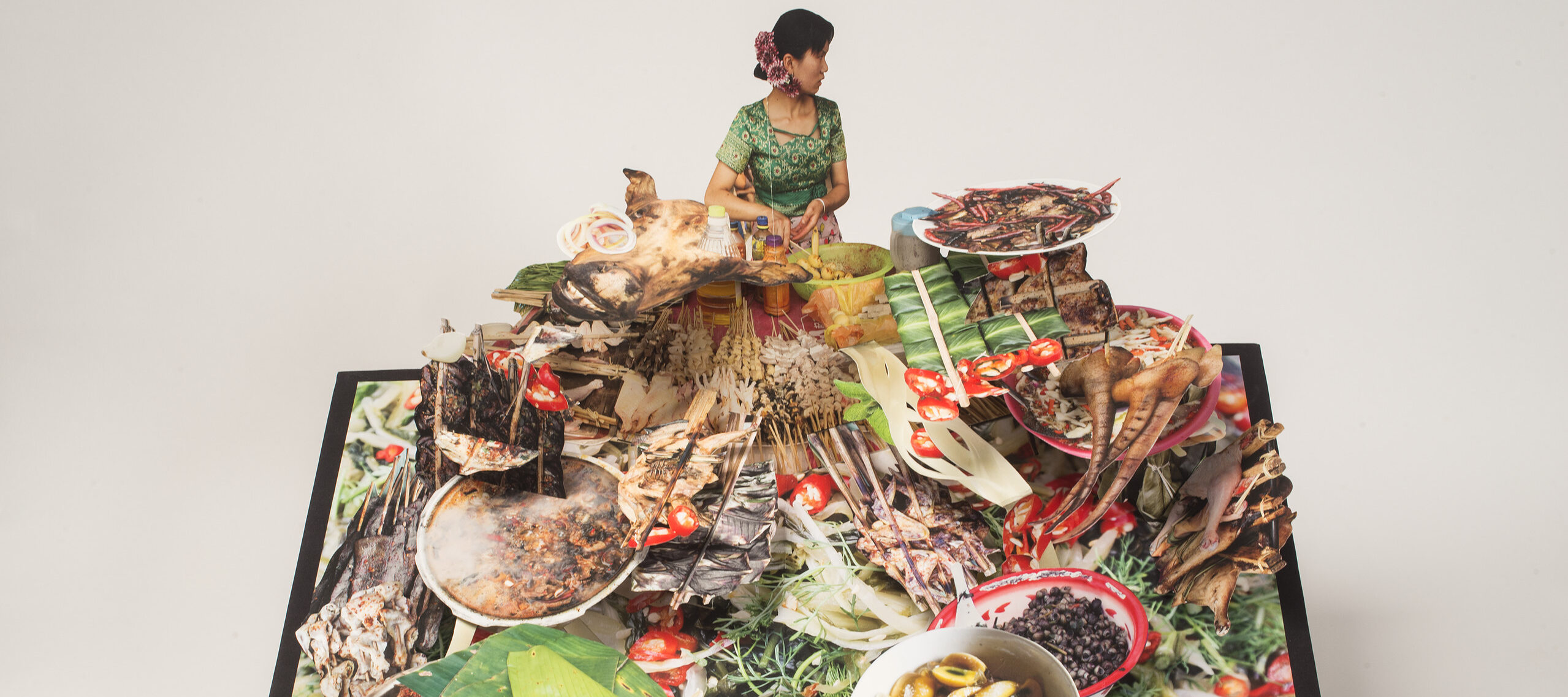 Large-scale pop-up book with a dark haired woman behind a huge display of food. She wears a green dress and purple flowers in her hair and turns to look over her left shoulder. The food explodes forward with bowls of soup, skewered meat, banana leaves, pig tail, and more.