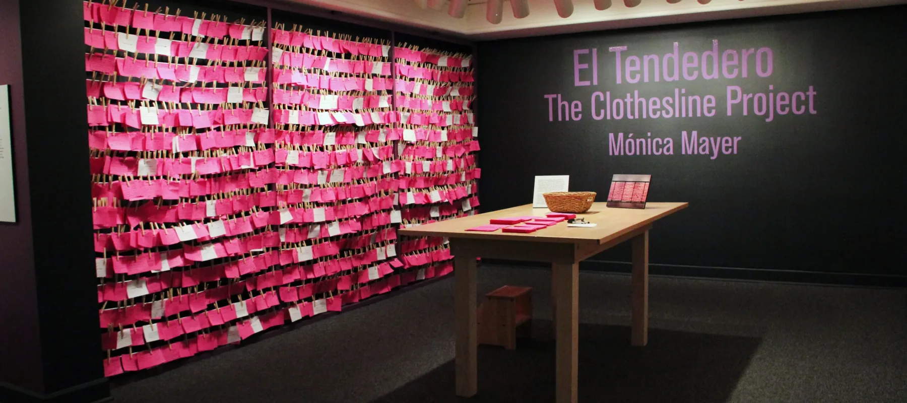 View of exhibition gallery features bright pink post-it notes line the wall from ceiling to floor. On the adjacent wall are the words in pink text:"El Tendedero, The Clothesline Project, Monica Meyer