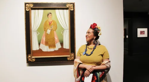 A medium skin toned woman in a yellow top and brightly patterned long skirt with flowers in her hair looks to her right at a painting of a similarly dressed woman.