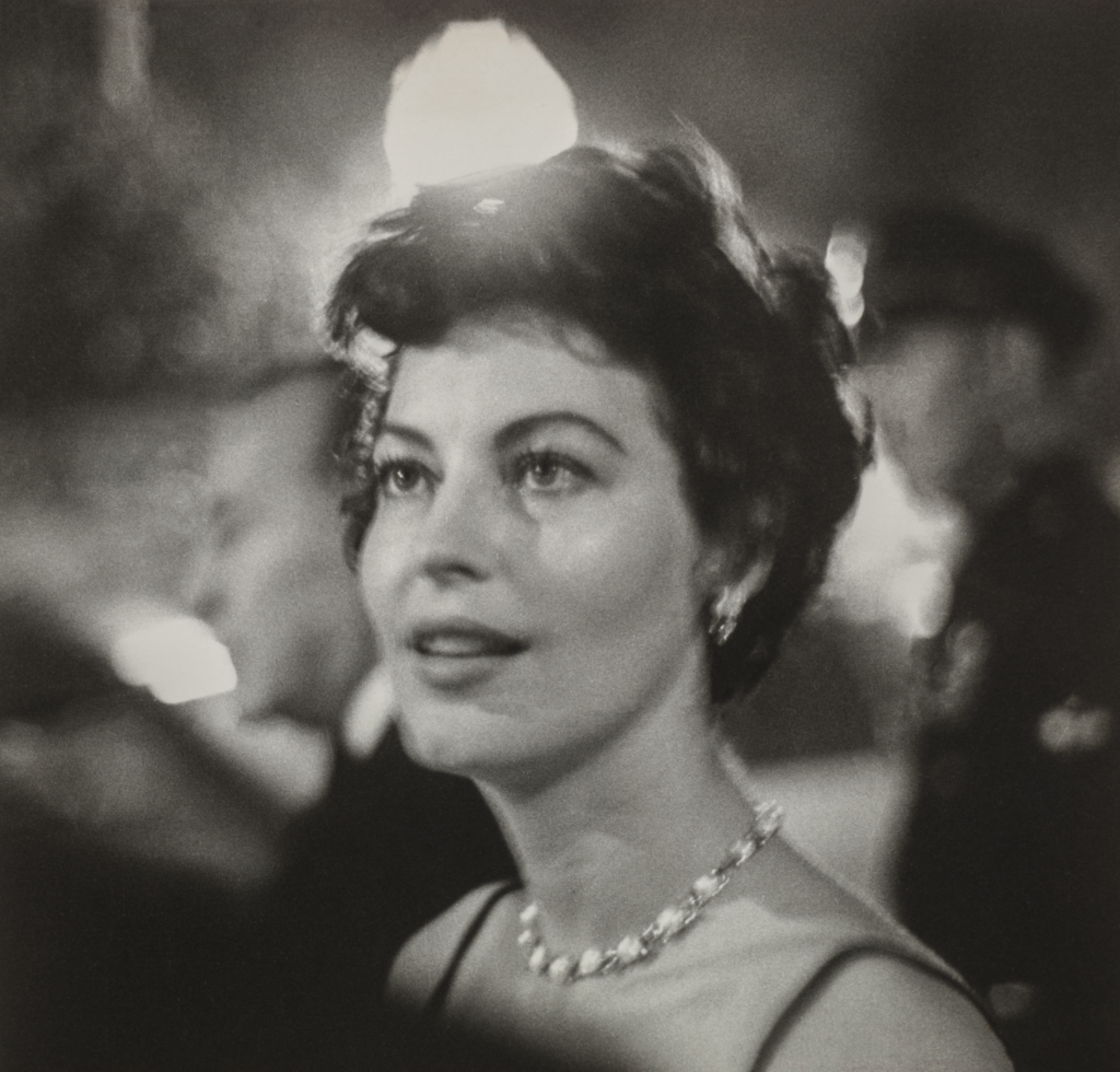 Ava Gardner, a woman with light skin tone and wavy, short brown hair, is shown from the shoulders up. She wears a jeweled necklace and is surrounded by lights and people at a party. She looks to the side of the camera at something out of frame with a fixed gaze and slight smile.