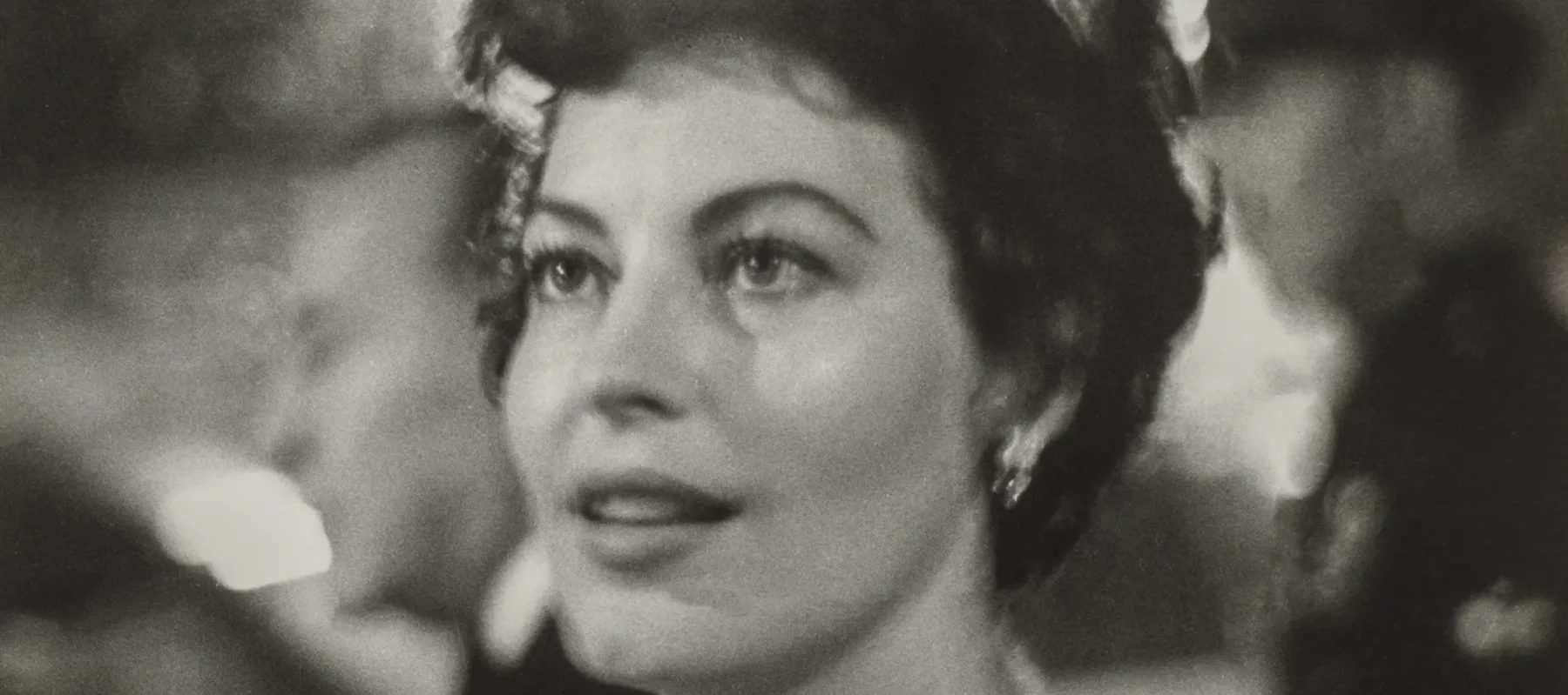 Ava Gardner, a woman with light skin tone and wavy, short brown hair, is shown from the shoulders up. She wears a jeweled necklace and is surrounded by lights and people at a party. She looks to the side of the camera at something out of frame with a fixed gaze and slight smile.