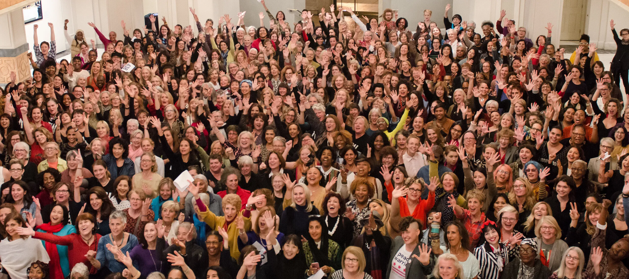 A color photograph of 435 women from a high vantage point. The women all look up towards the camera, some with arms outstretched.