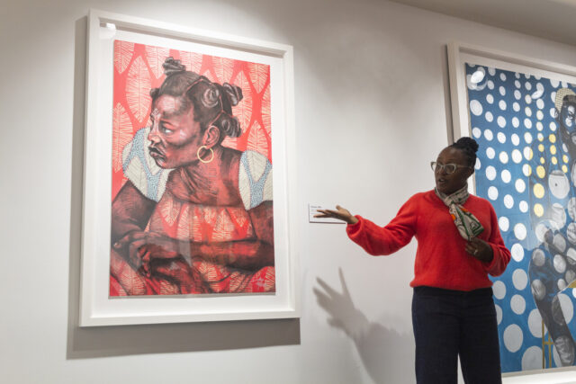 The artist, a woman with dark skin tone, speaks in front of her artwork, gesturing to it, a large colorful print of a woman with a pattern overlay across the paper. The artist wears a bright right sweater, a multicolored scarf, glasses, and gold hoops.