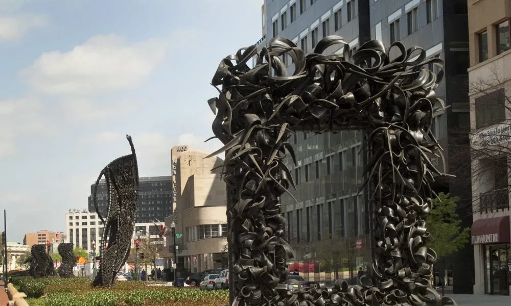 A large metal sculpture stands in the middle of the street, similar to an empty rectangular picture frame. The “frame” is crowded with undulating, coiling metal sculptural elements that occasionally end in spikes. Behind stand three vertical twisting, net-like metal sculptures.