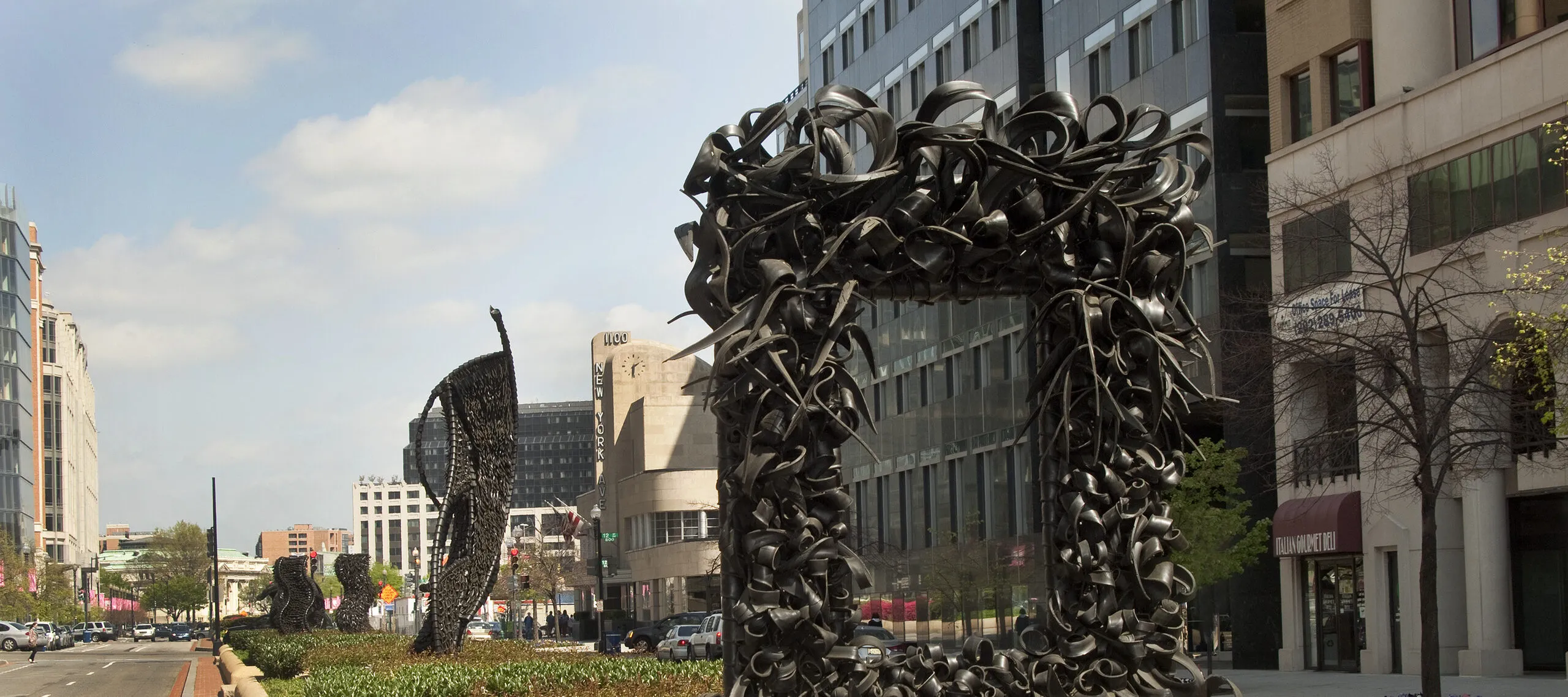 A large metal sculpture stands in the middle of the street, similar to an empty rectangular picture frame. The “frame” is crowded with undulating, coiling metal sculptural elements that occasionally end in spikes. Behind stand three vertical twisting, net-like metal sculptures.