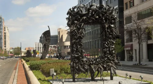 A large metal sculpture stands in the middle of the street, similar to an empty rectangular picture frame. The “frame” is crowded with undulating, coiling metal sculptural elements that occasionally end in spikes. Behind stand three vertical twisting, net-like metal sculptures.