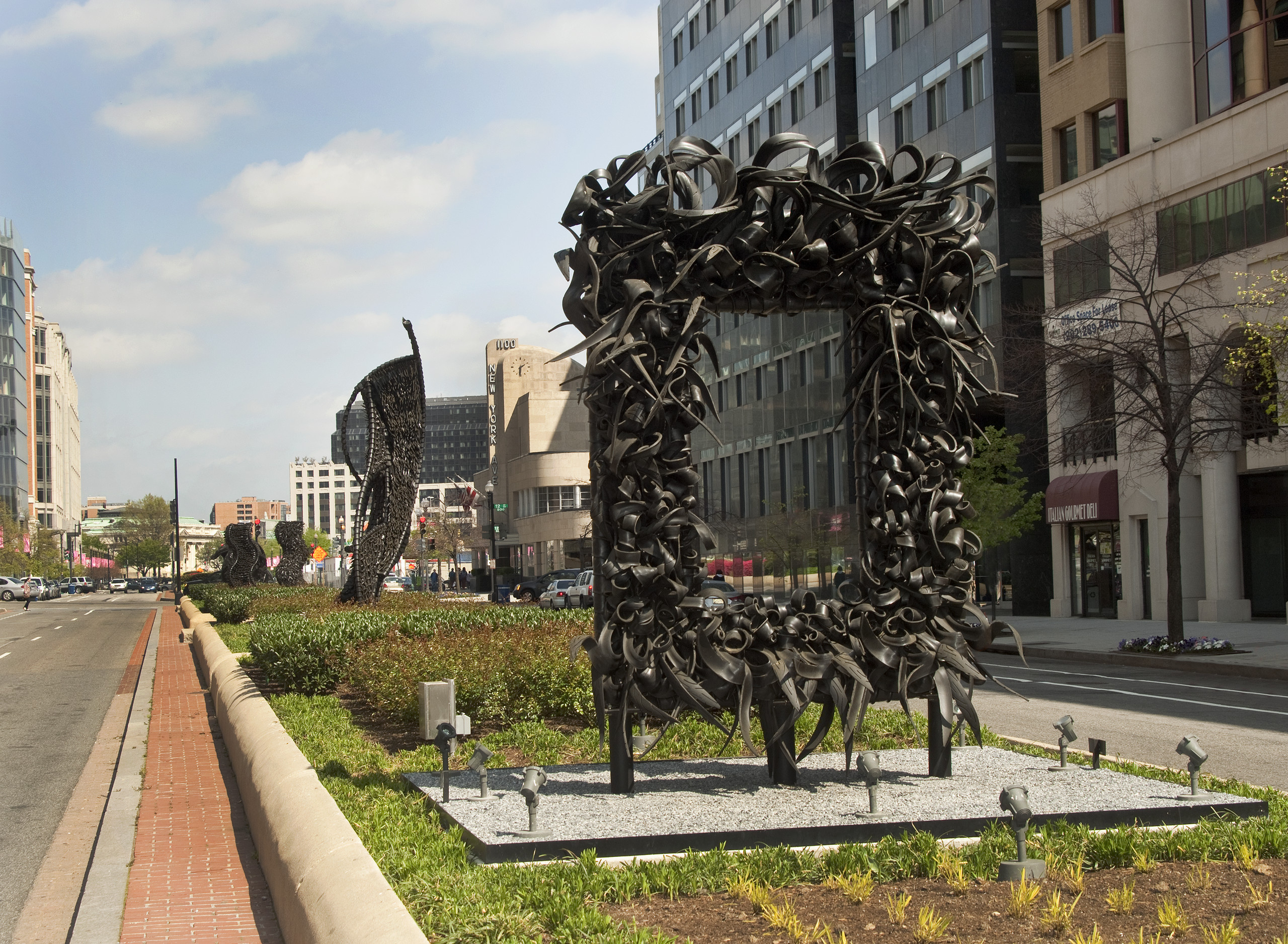 A large metal sculpture stands in the middle of the street, similar to an empty rectangular picture frame. The “frame” is crowded with undulating, coiling metal sculptural elements that occasionally end in spikes. Behind stand three vertical twisting, net-like metal sculptures.
