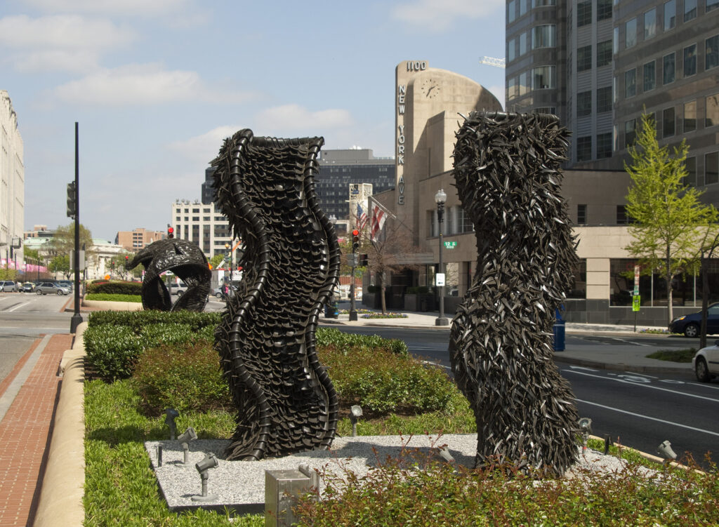 Two large abstract sculptures made from tires rise from a center median in a city street. The sculptures are wavy columns. 