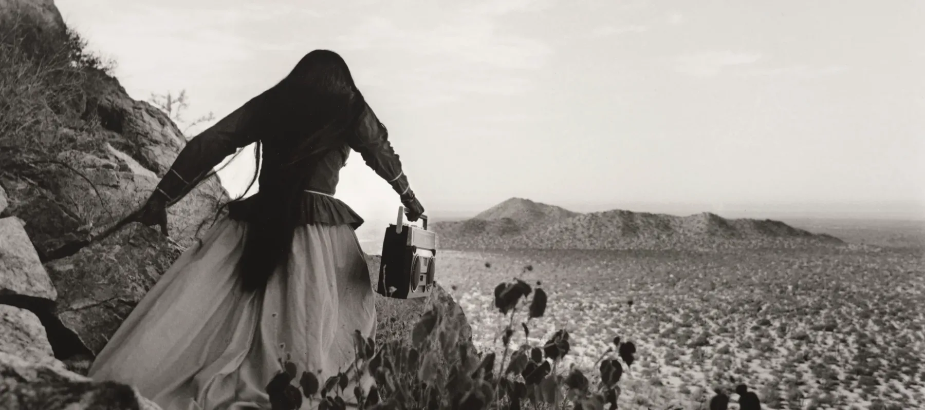 A black-and-white photograph shows the back of a woman as she crests a rocky path above a vast desert landscape beneath an expansive sky. Her traditional, ethnic full skirt, long-sleeved blouse, and long, straight, dark hair contrasts with the modern portable stereo she carries.