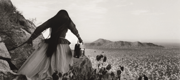 A black-and-white photograph shows the back of a woman as she crests a rocky path above a vast desert landscape beneath an expansive sky. Her traditional, ethnic full skirt, long-sleeved blouse, and long, straight, dark hair contrasts with the modern portable stereo she carries.