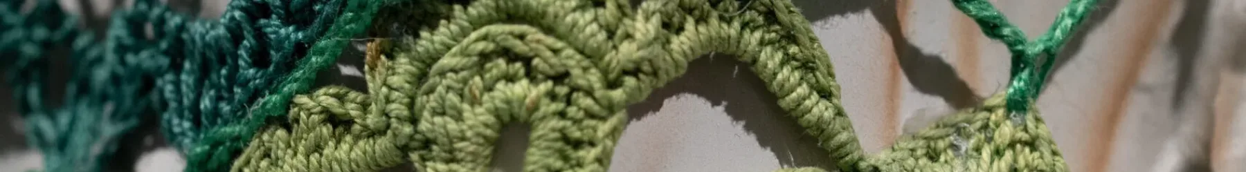 Close-up detail of a larger artwork shows an off-white ceramic surface that is covered by panels of crocheted yarn in emerald and citron green.