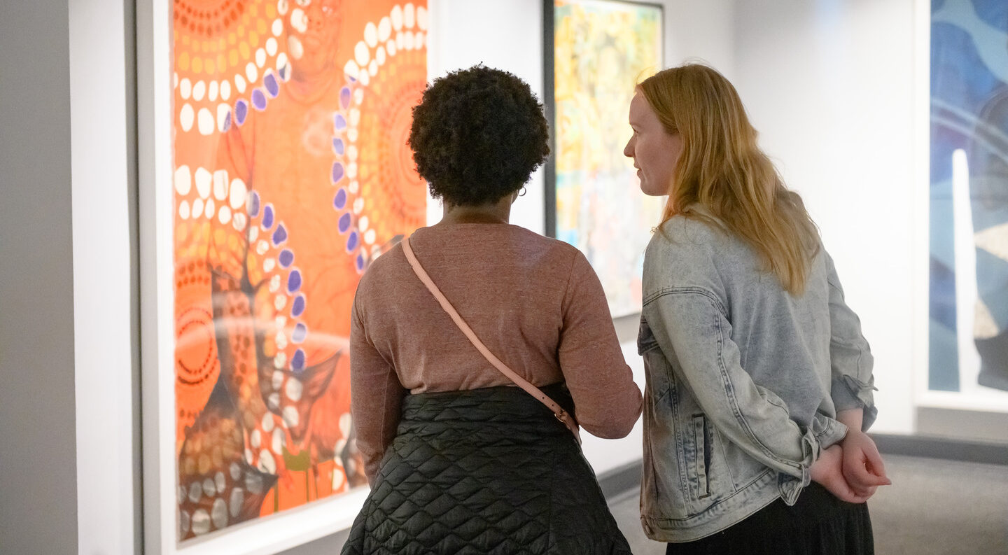 Two figures stand facing a large colorful work on paper. The figure on the left is dark skinned with short, curly, brown hair, the figure on the right is light skinned with long, straight, blonde hair. The work on paper depicts a seated dark skin person staring confidently at the viewer; an overlay of bright orange and circular patterns of white, blue, and brown circles covers the person and background.