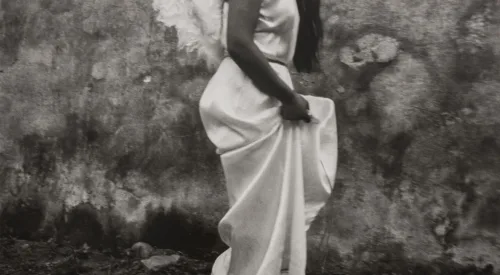 A black-and-white photograph of a woman in profile from the shoulders down with feathery white wings on her back walking and hoisting up her silky white dress.