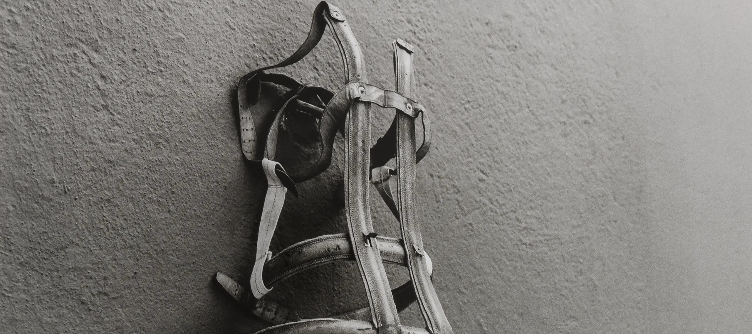 A black and white photograph of a worn body brace pinned on a blank concrete wall. The photograph is taken from slightly below and to the left of the brace. Similar to a corset, the brace itself is simultaneously lonely, foreboding, and empty as it hangs.