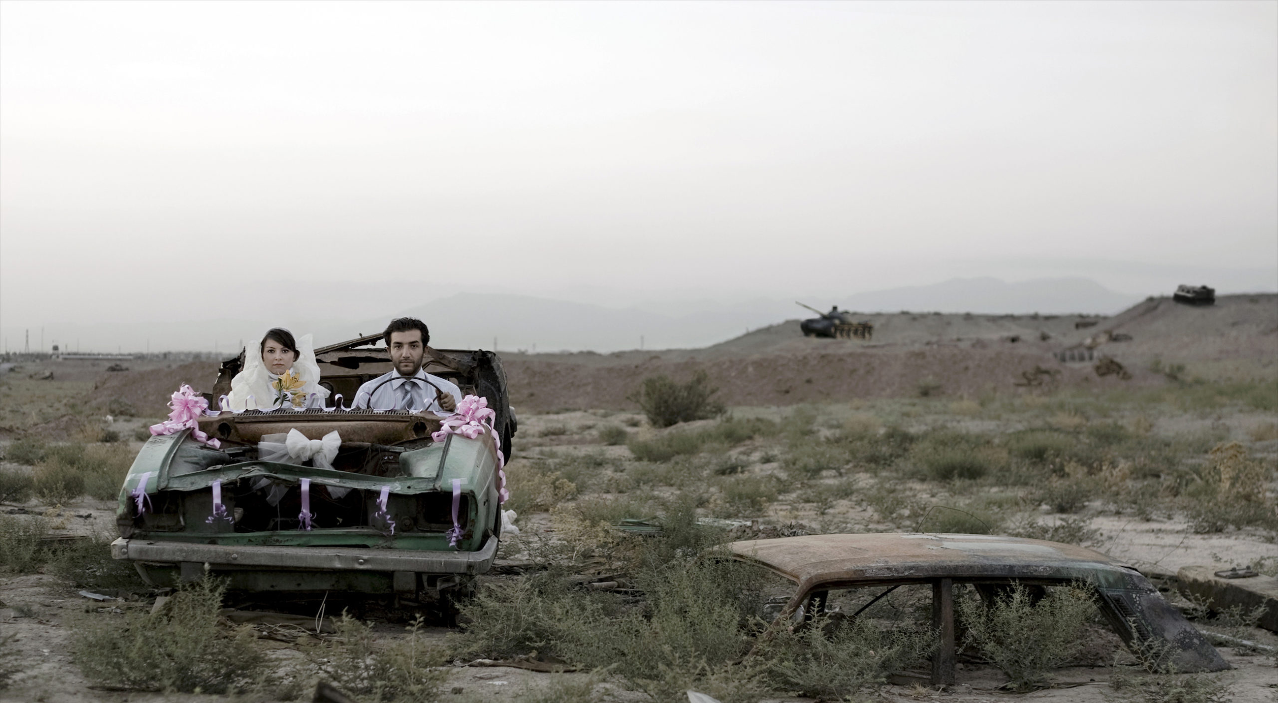 A couple sitting in a burned-out car in wedding finery, they look directly at the camera with neutral or stricken expressions.