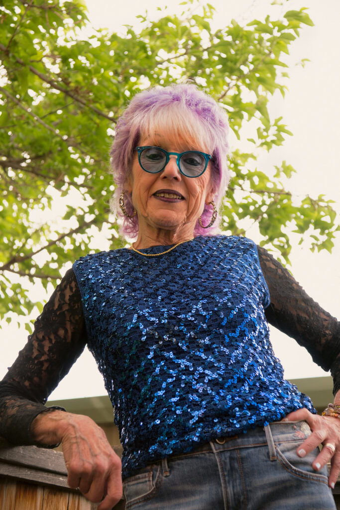 A light-skinned older woman with short purple-and-white hair smiles slightly, arms out to the side. She wears a blue sequin top with long black lace sleeves, a gold necklace, hoop earrings, and blue glasses. The sky and tree branches are behind her.