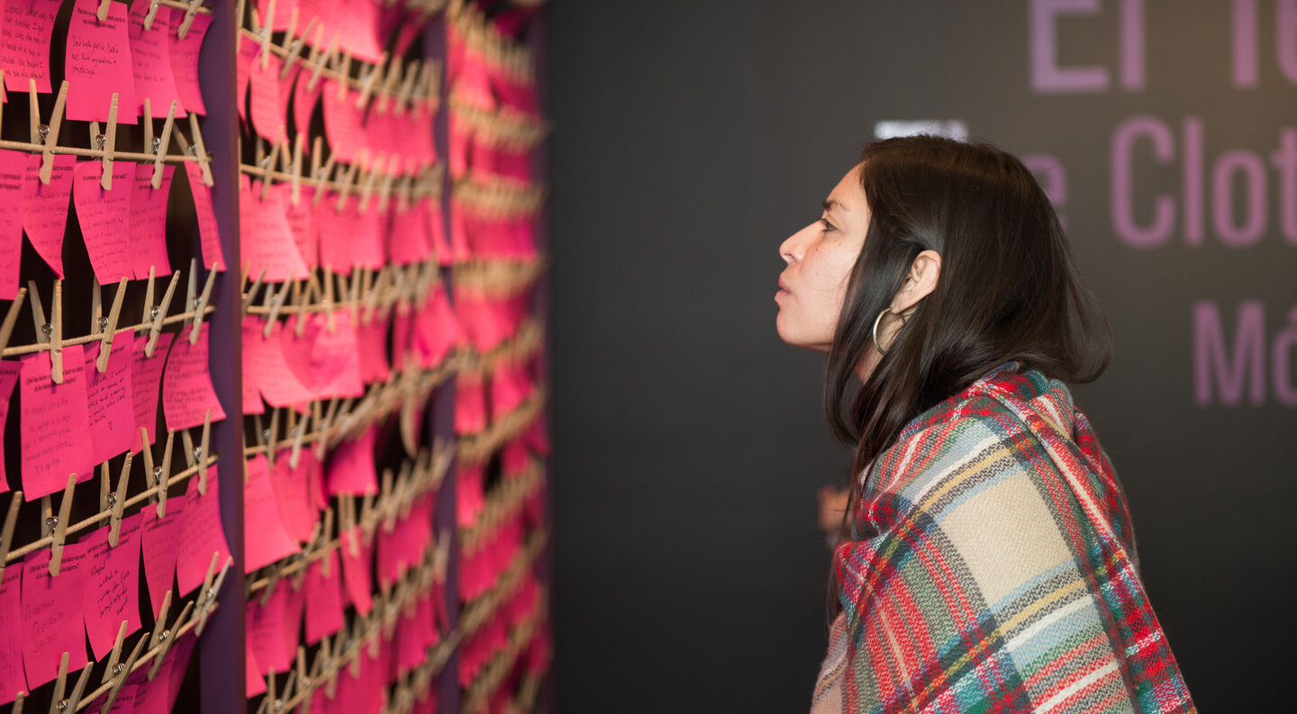 A light-skinned woman with dark hair looks at a wall of bright pink notes hung on multiple rows.