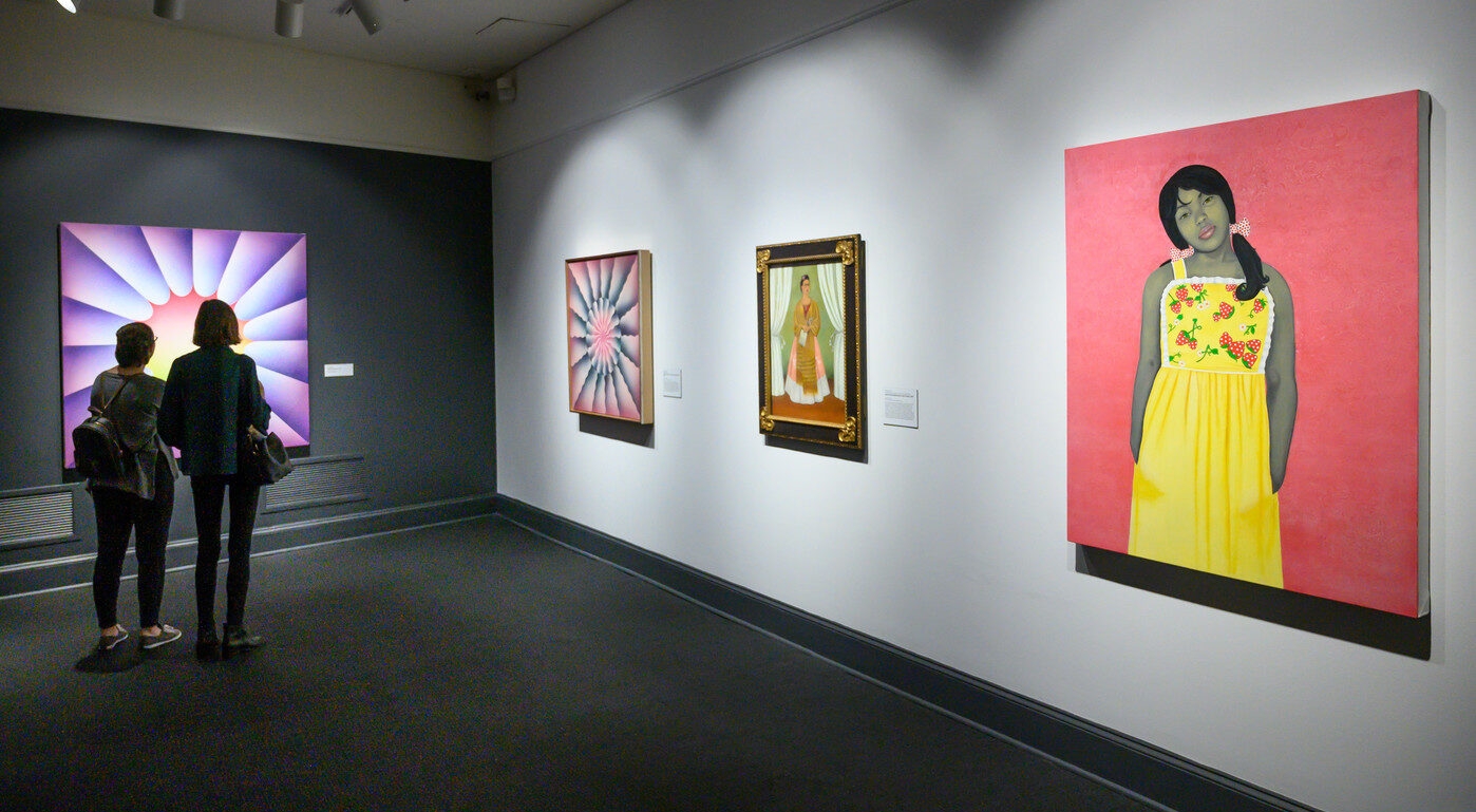 A partial view of an art gallery with four artworks on display, including a painted portrait of a young, pony-tailed medium-dark skinned girl in a yellow flowered dress against a pink background; a smaller portrait; and large abstract works with colorful shapes and gradients.