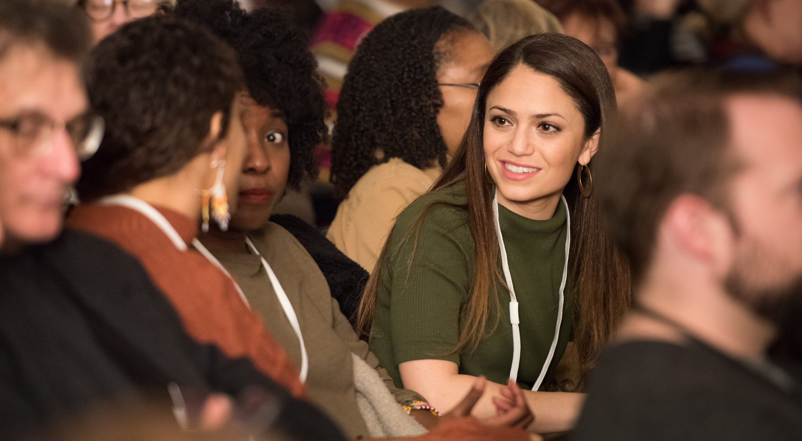A medium light-skinned young adult smiles while listening to another adult speak a few seats over from her in a full auditorium.