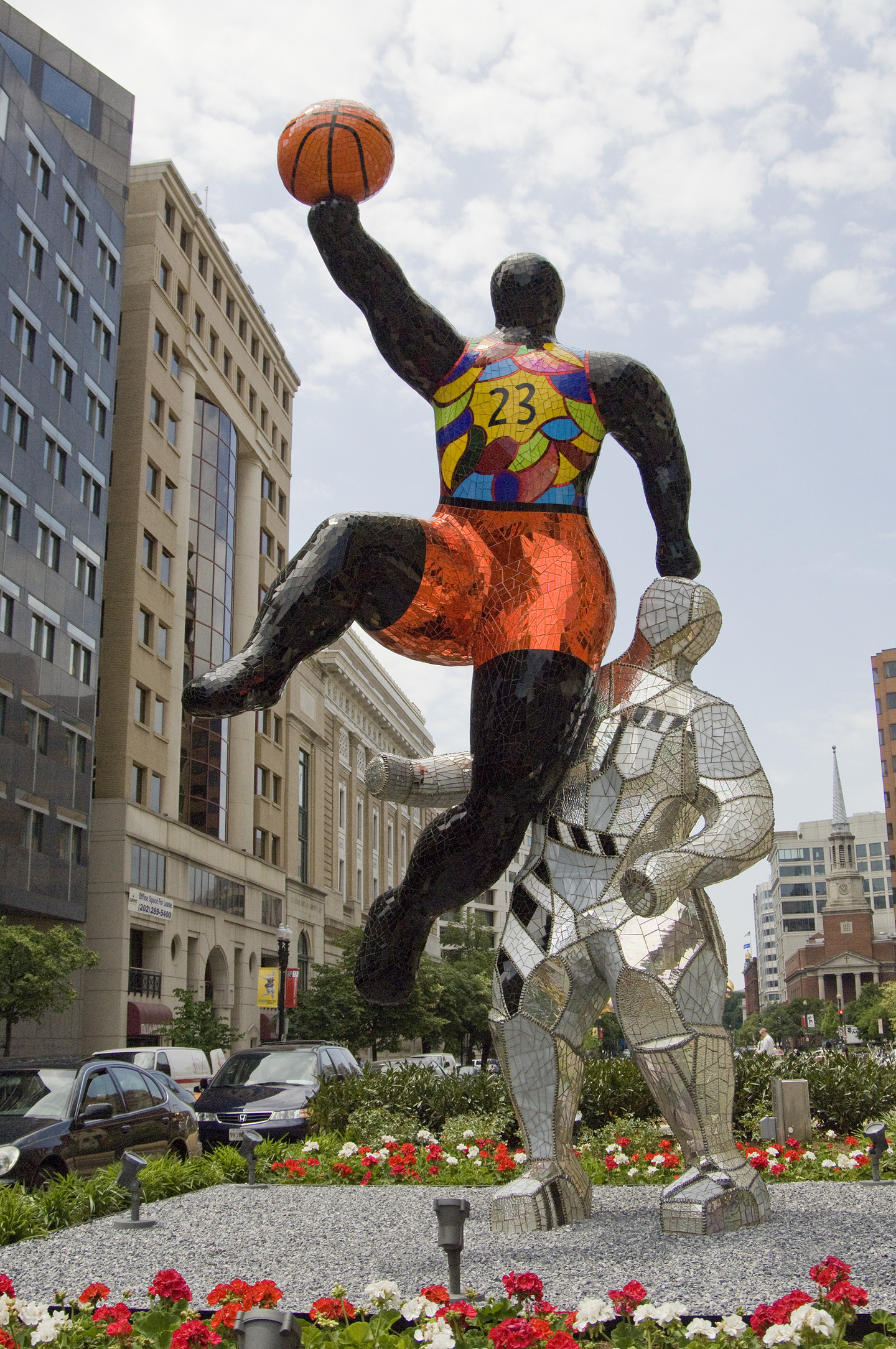Two large figures, one black and one in silver, are playing basketball next to a street. The figure with the black skin is jumping up with the basketball, leaning their hand onto the head of the figure in silver. The figure jumping is wearing a colorful outfit made from a reflecting material.