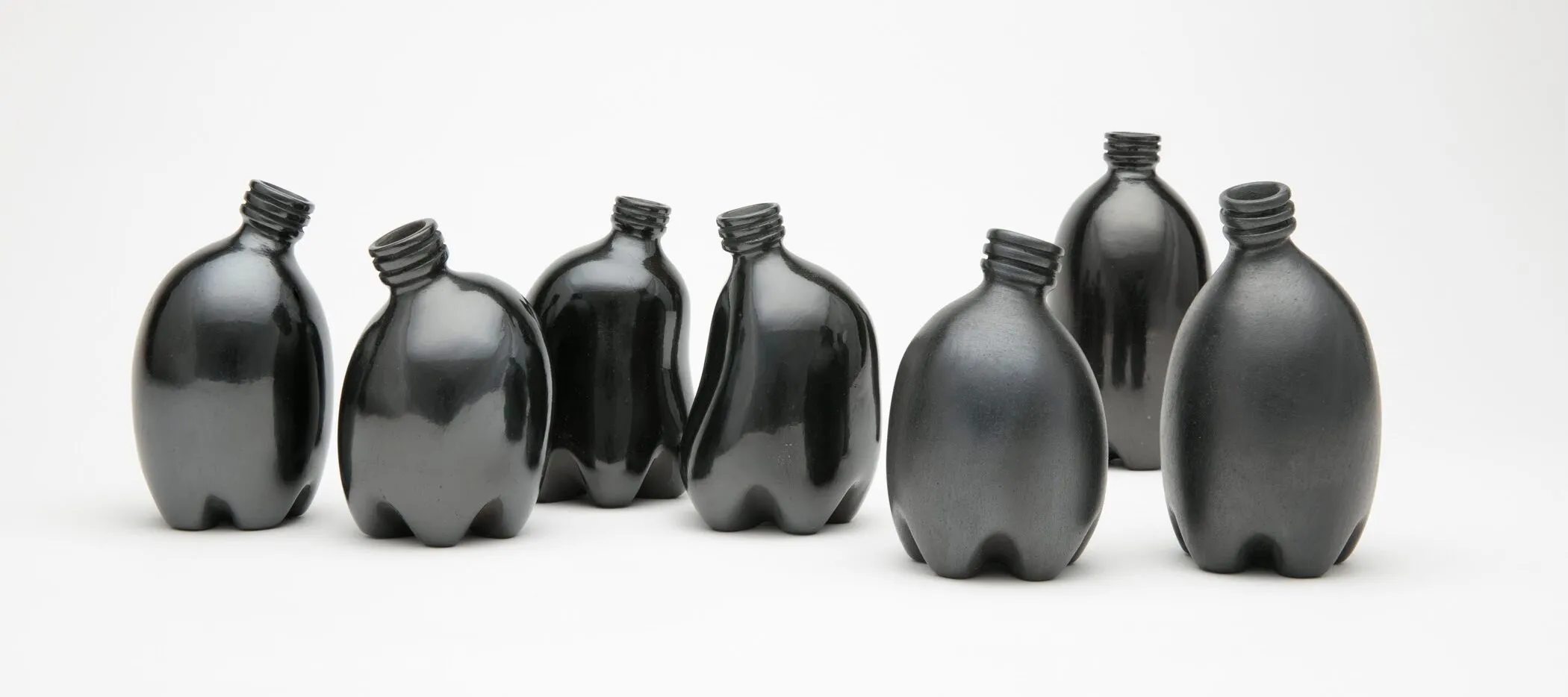 Against a stark white background are seven small clay vessels painted a dark, shiny black. They look like miniature plastic water bottles and are each slightly bent, making them appear like a gathering of people interacting.