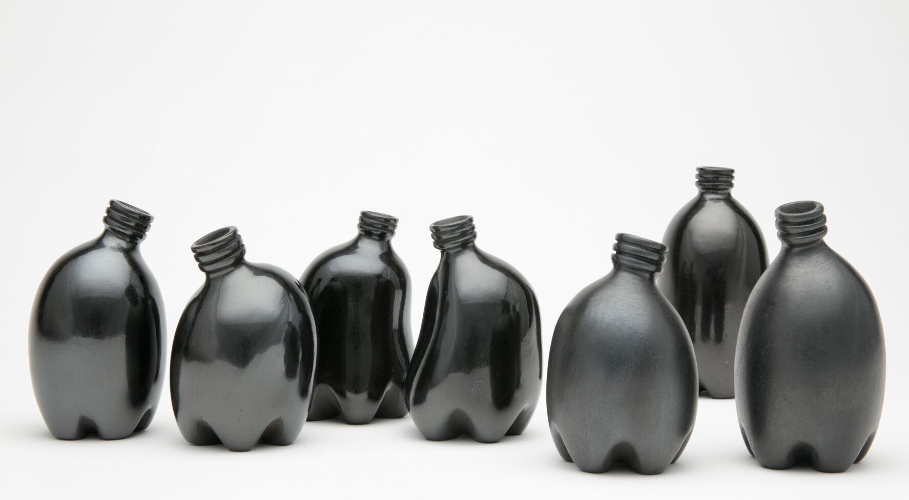 Against a stark white background are seven small clay vessels painted a dark, shiny black. They look like miniature plastic water bottles and are each slightly bent, making them appear like a gathering of people interacting.