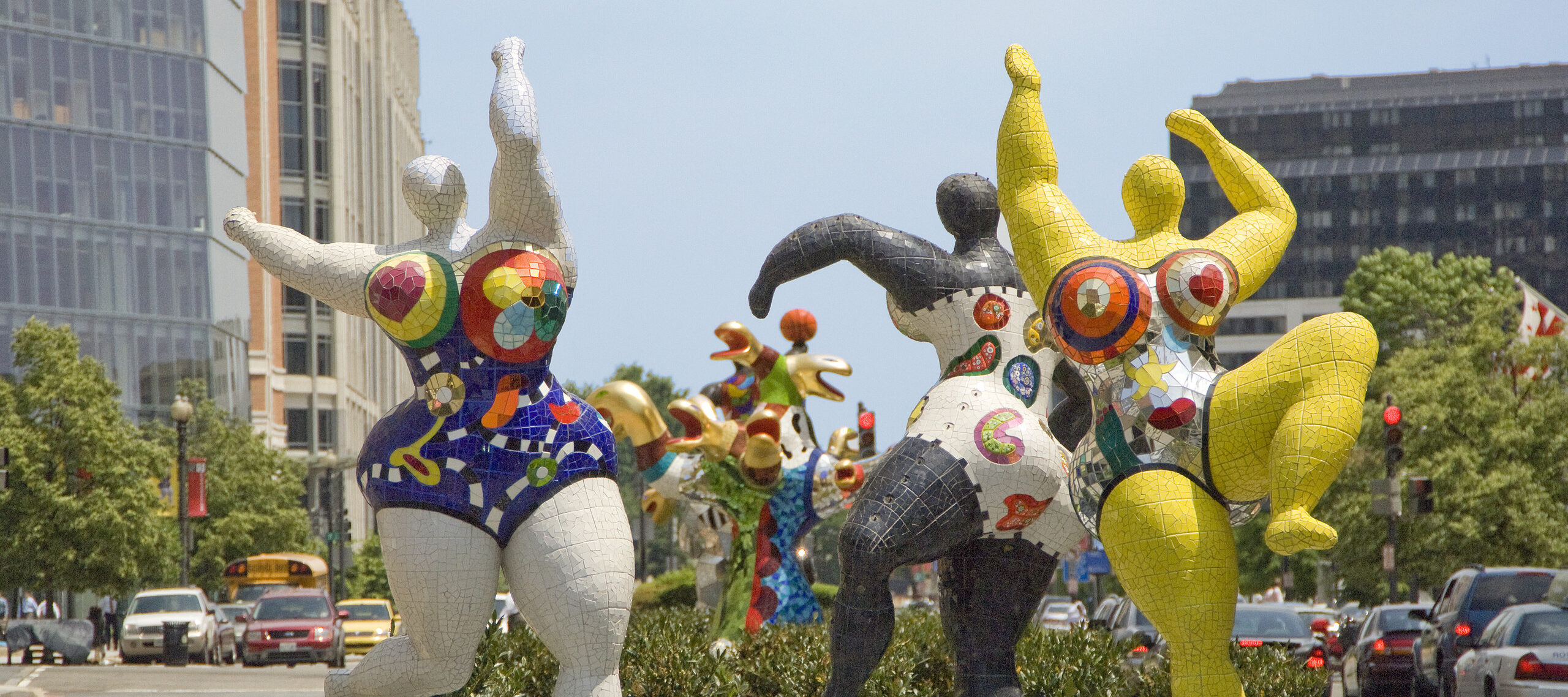 Three large figures are dancing along a street. The figures have no faces but womanly bodies. They are wearing colorful bikinis with hearts and flowers painted on them. The figures appear enormous compared to the cars standing nearby.