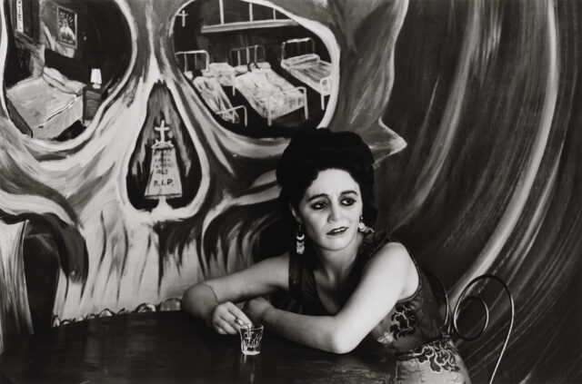 Black and white photograph of light skin toned woman with long dark hair sitting at a cafe table. On the wall behind her is a painted mural of a large skull with hospital beds visible in the eye sockets and a tombstone in the nasal cavity.