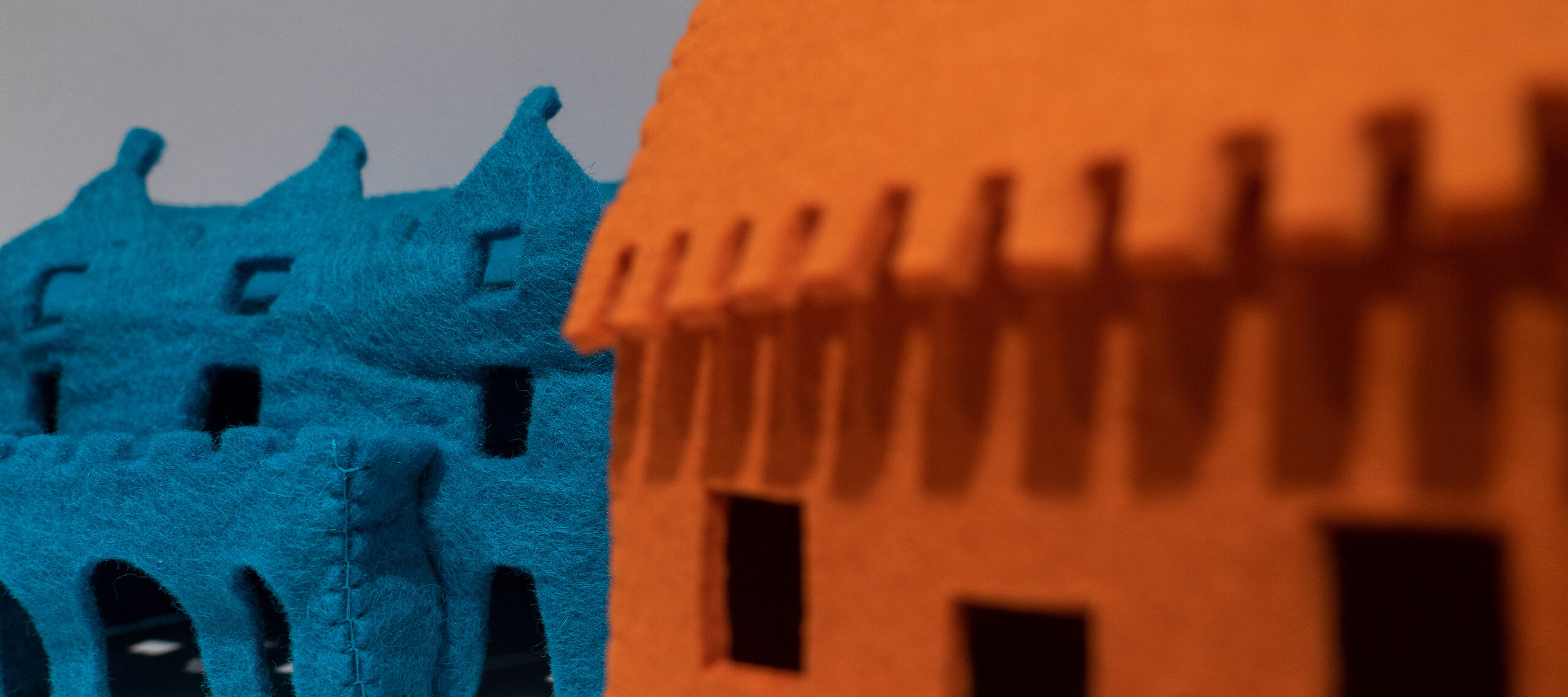 Close-up detail of a larger artwork features a view of two felt houses. On the viewer's right is an orange house that is out-of-focus while on the left is a blue house that is in-focus and has visible stitches, coarse texture, and rectangular windows.