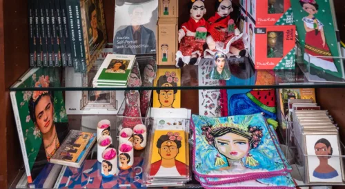 Three clear glass shelves, packed with colorful products on display. The top two shelves are full of Frida Kahlo-themed items—books, dolls, cards, and more. The bottom shelf is full of Louise Bourgeois-themed items—teapots, a sleep mask, books, mugs, and more.