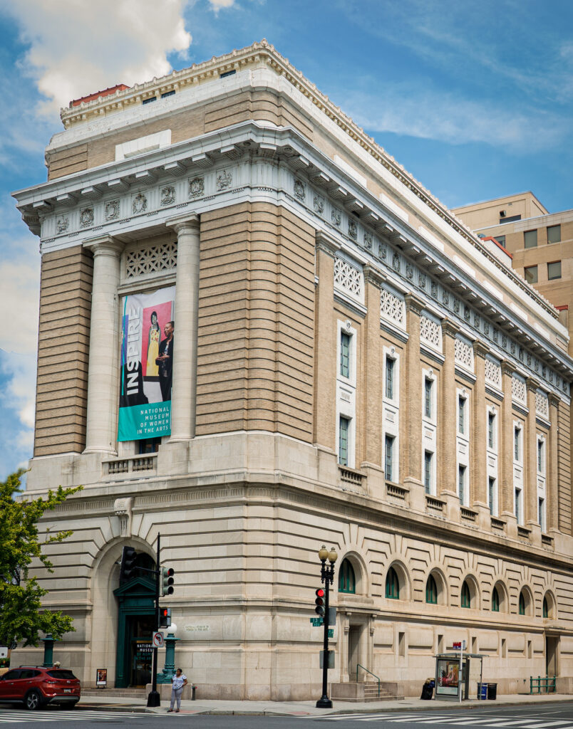 View of the museum from outside showing the Neoclassical building from one corner. The building is a tan-colored stone with an arched doorway, long vertical windows, and detailed molding around the roof.