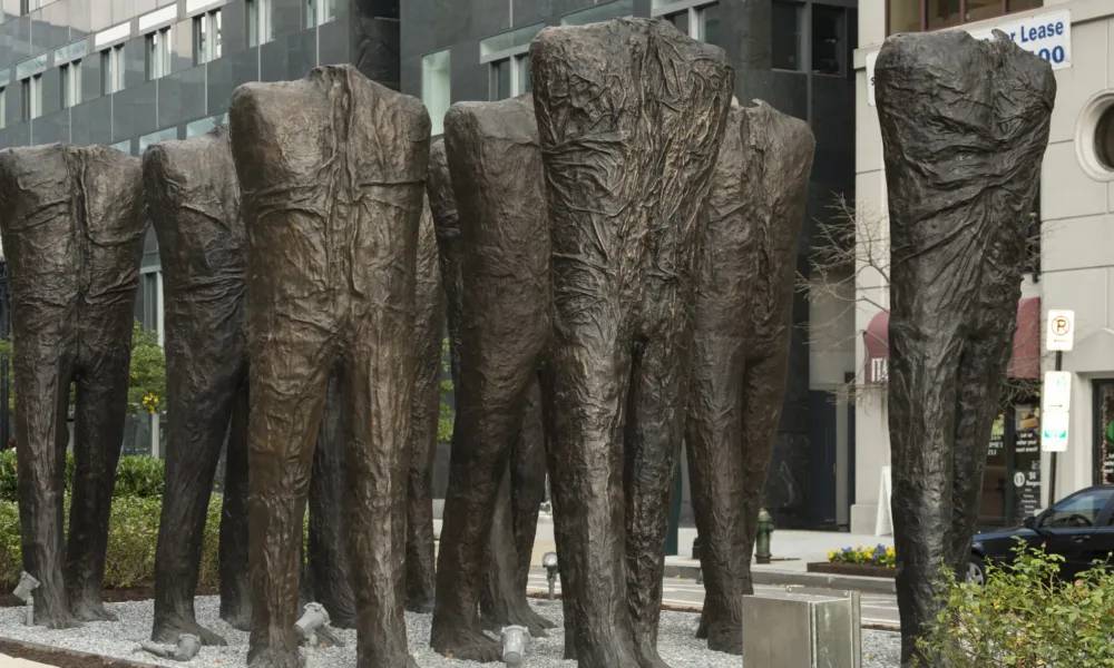 Ten larger-than-life bronze sculptures of human bodies are installed in the middle of a city street. The bodies have no heads or arms, and are striding forward in five rows of two. While they are not naked, their wrinkled body-tight clothing makes no distinction between shirt and pants.