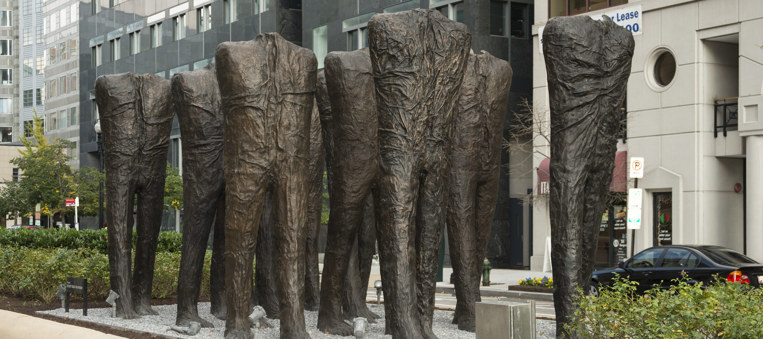 Ten larger-than-life bronze sculptures of human bodies are installed in the middle of a city street. The bodies have no heads or arms, and are striding forward in five rows of two. While they are not naked, their wrinkled body-tight clothing makes no distinction between shirt and pants.