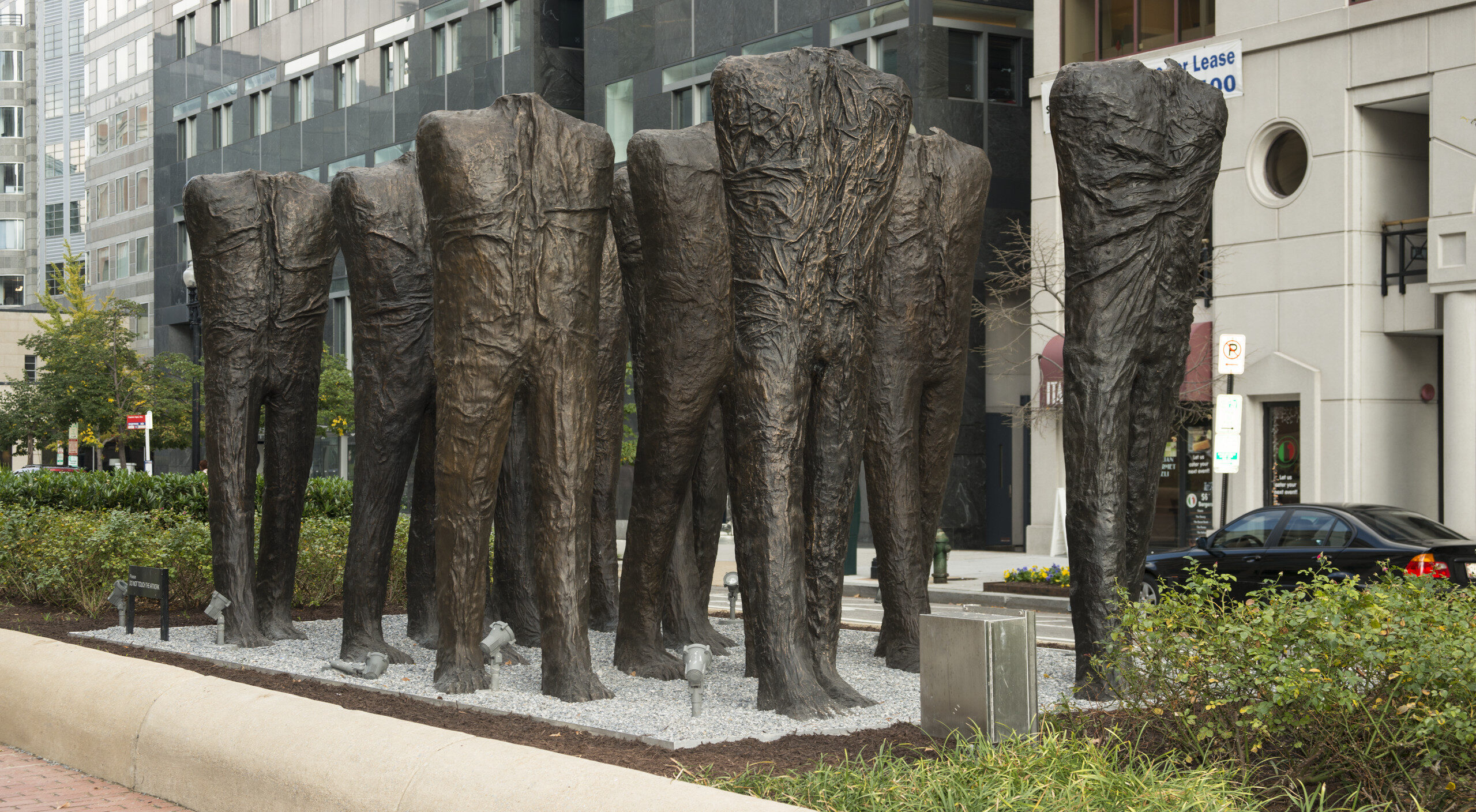 Ten larger-than-life bronze sculptures of human bodies are installed in the middle of a city street. The bodies have no heads or arms, and are striding forward in five rows of two. While they are not naked, their wrinkled body-tight clothing makes no distinction between shirt and pants.