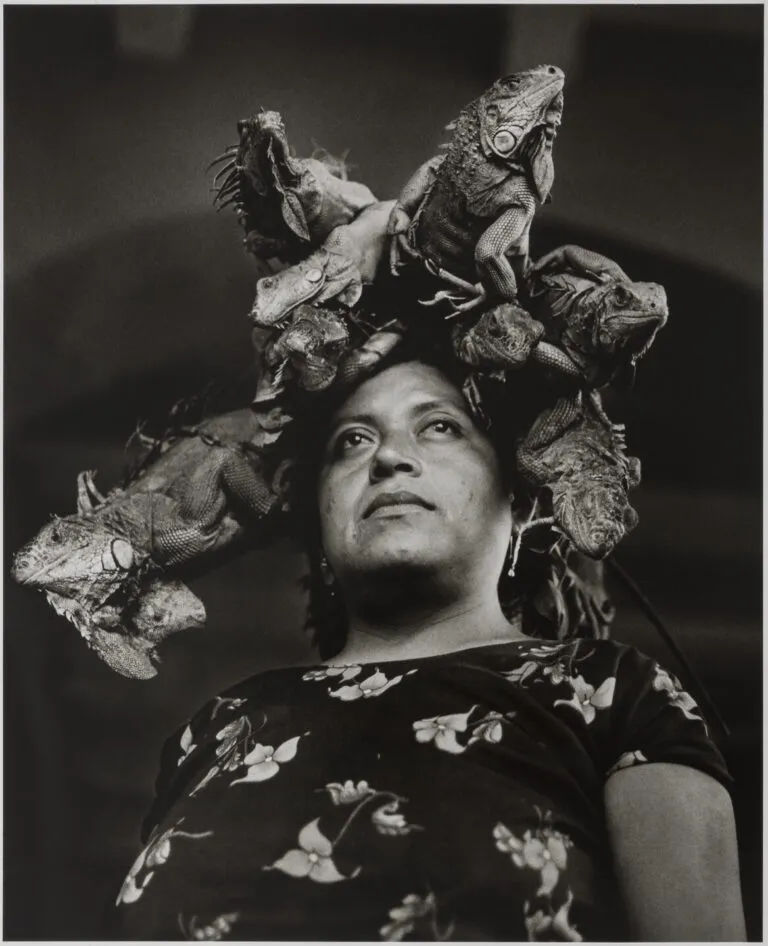 Black and white photograph of a Mexican woman looking off into the distance. She wears a floral blouse and five iguanas sit atop her head at different angles, as if she were wearing a crown.