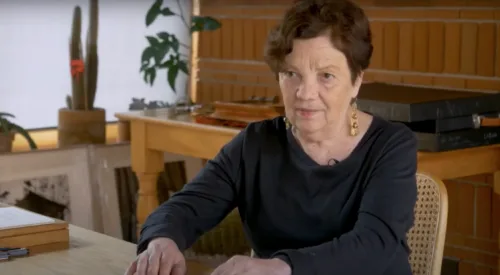 Still photograph from a video about the artist shows an older woman with light skin and dark hair sitting at a table.