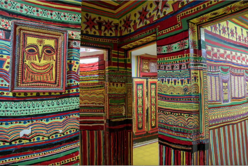 An interior photograph of a home. The walls are mostly red, yellow, green, and blue; vertical stripes ascend the walls until waist height, where horizontal registers of stripes, dots, and scalloped patterns take over. The upper third of the walls are painted in varied repeating shapes like stars and triangles.