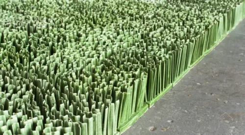 Detail photograph of ceramic sculpture made to look like a patch of lawn. Individual squares consisting of multiple upright blades of porcelain grass, glazed green, fit together to form a lush rectangular field of grass.