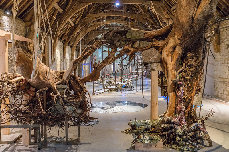 Various elements of an art installation are housed in a cavernous room with a vaulted wood ceilings and stone walls. In the front, two large trees grow towards each other. They have no foliage, and instead display networks of roots, which are painted in patterns and colors.