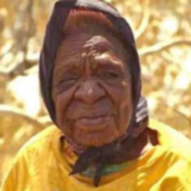 An older woman with dark skin tone wears an eggplant-colored scarf around her head and a yellow shirt squinting at the camera.