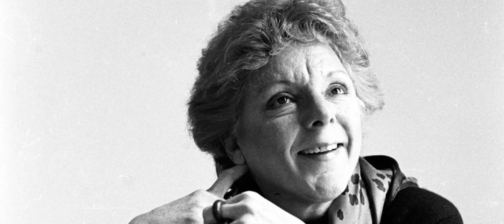 A black and white photographic portrait of seated art scholar Linda Nochlin. Dressed in a dark sweater and a print scarf, she gazes with a smile up to her left. An oversized art book is opened on the surface in front of her.