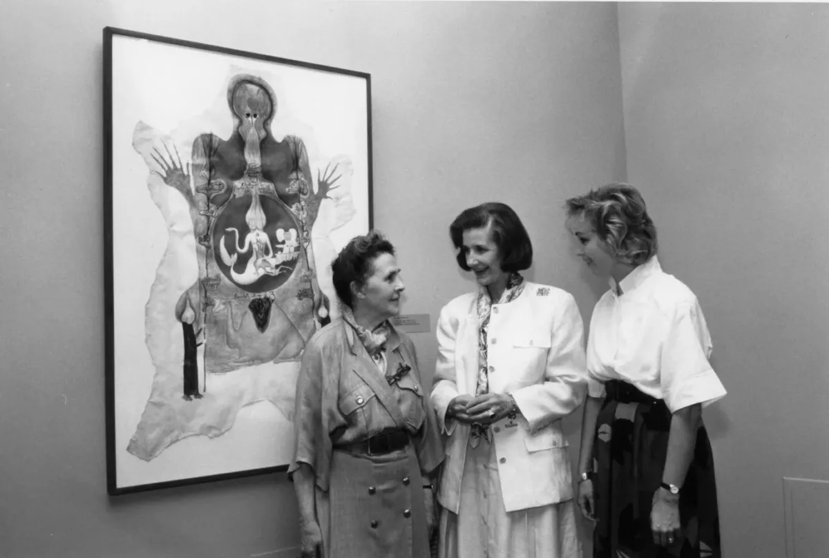 Black and white photograph of three light skinned women casually chatting next to a painting.