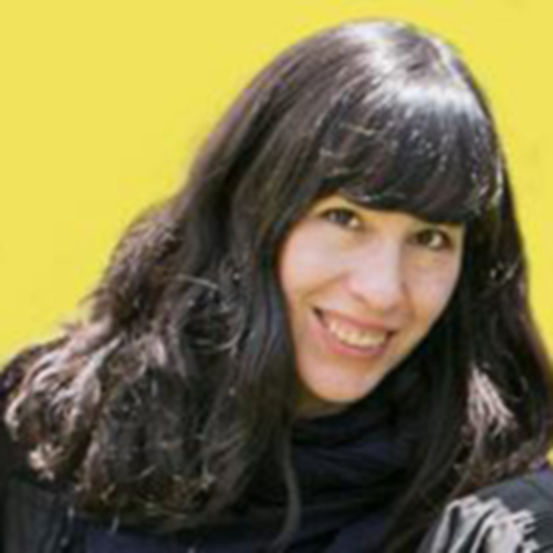 A photograph of an adult woman long, straight, dark hair and thick bangs covering her forehead. She is posed from the neck up against a bright yellow background. She makes eye contact with the viewer and is smiling.