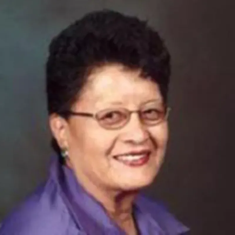 Headshot of a middle aged woman with medium-light skin tone, short dark hair, glasses, earrings, and a purple top.