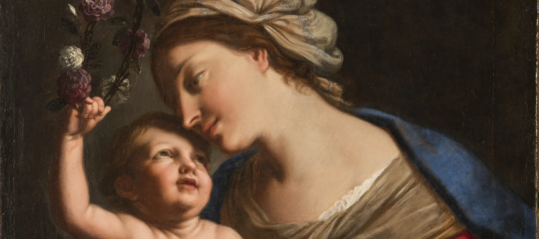 A young, light-skinned, brunette woman gazes down lovingly at the plump baby she holds on her lap. She wears a loose, tan turban, vivid blue cloak, and red dress with white sleeves. The light-skinned child returns her gaze, leaning back to crown the woman with a circlet of roses.