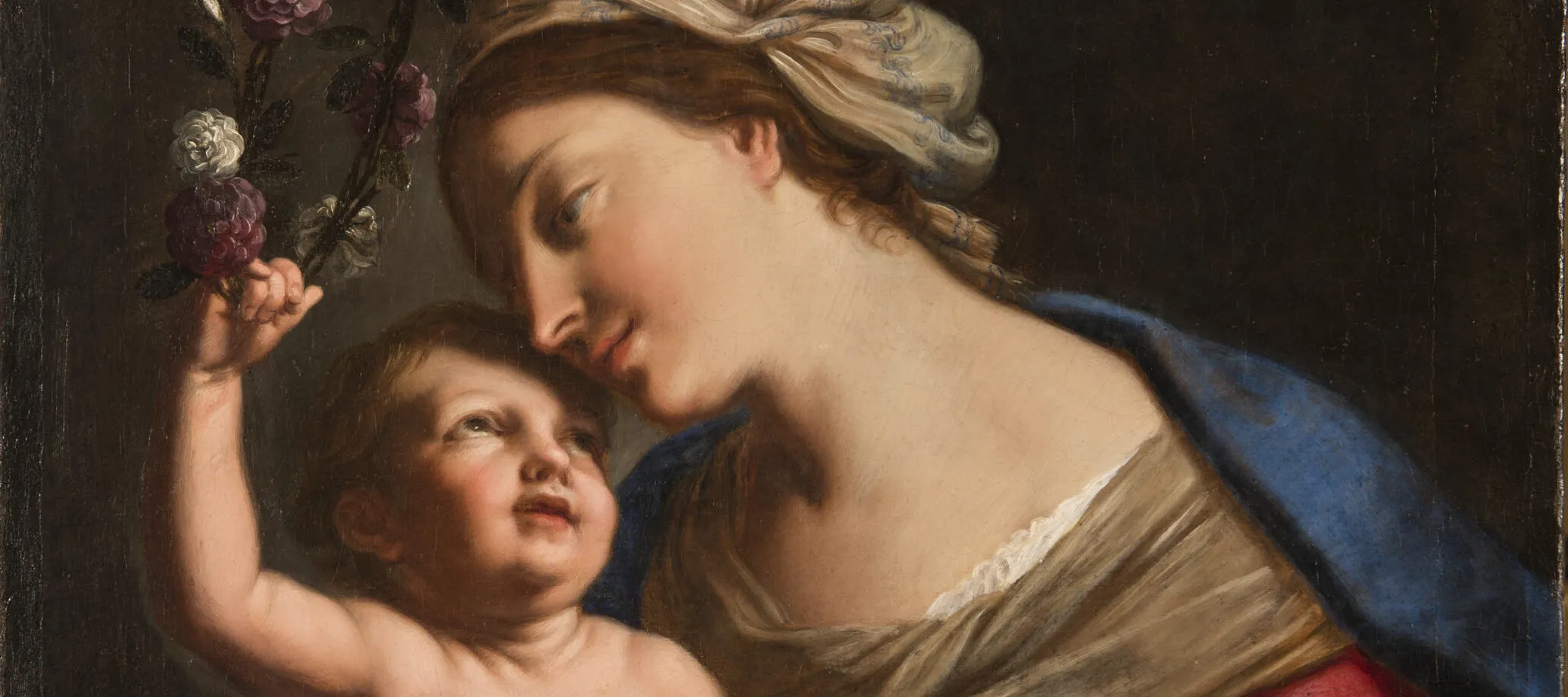 A young, light-skinned, brunette woman gazes down lovingly at the plump baby she holds on her lap. She wears a loose, tan turban, vivid blue cloak, and red dress with white sleeves. The light-skinned child returns her gaze, leaning back to crown the woman with a circlet of roses.