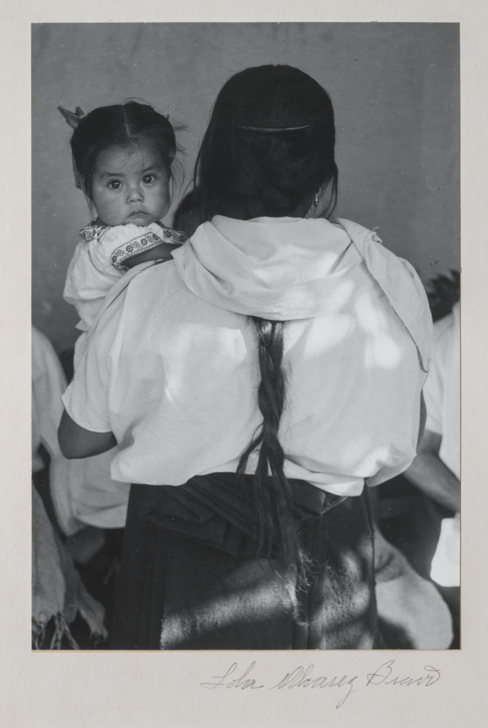 Black-and-white photograph of a woman wearing a dark skirt and light blouse holding a child. The woman faces back and her dark braid reaches past her waist. The dark-haired child stares at the camera and wears a white embroidered dress. Other figures are slightly visible beyond.