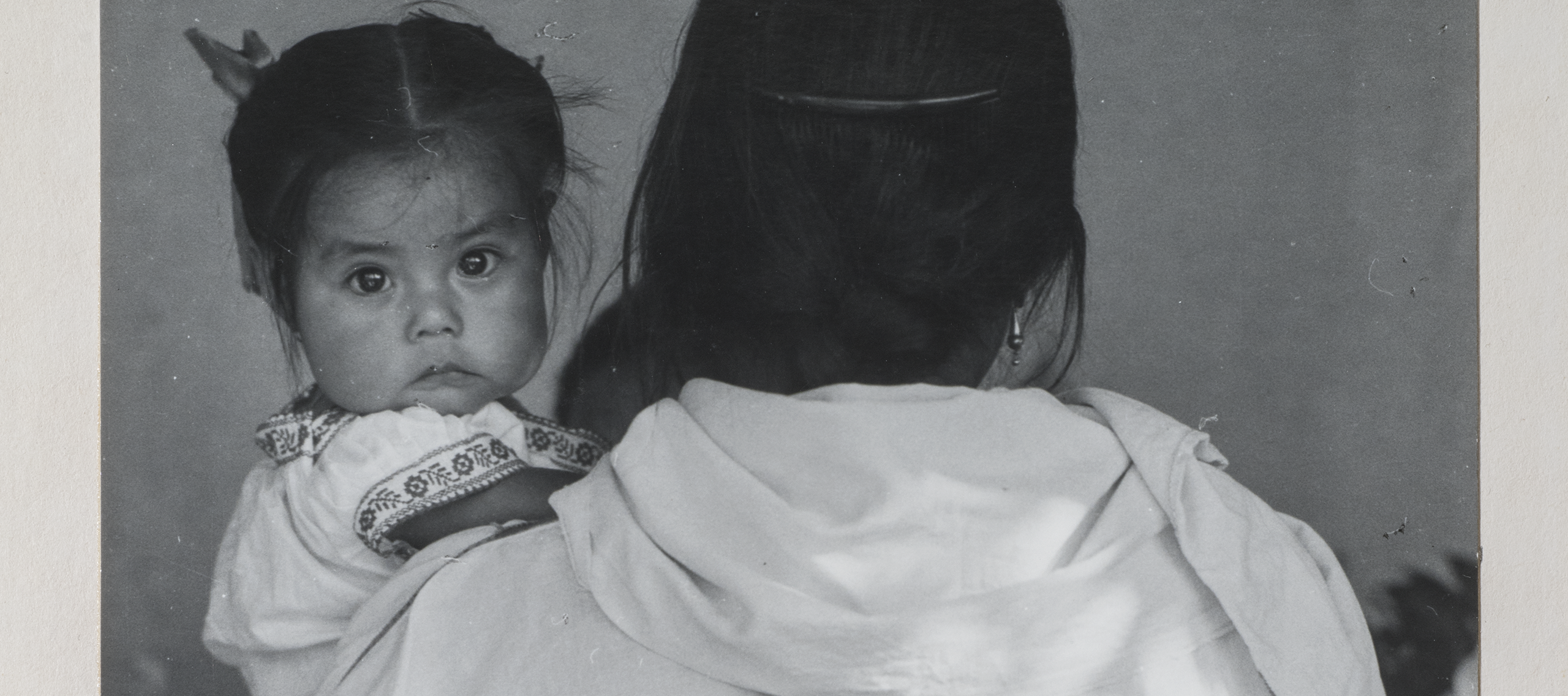 Black-and-white photograph of a woman wearing a dark skirt and light blouse holding a child. The woman faces back and her dark braid reaches past her waist. The dark-haired child stares at the camera and wears a white embroidered dress. Other figures are slightly visible beyond.