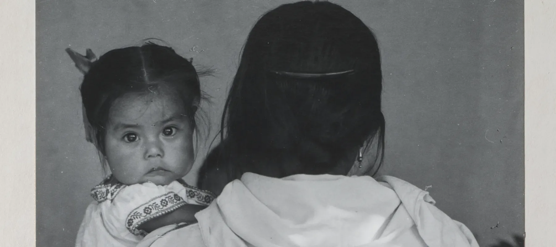 Black-and-white photograph of a woman wearing a dark skirt and light blouse holding a child. The woman faces back and her dark braid reaches past her waist. The dark-haired child stares at the camera and wears a white embroidered dress. Other figures are slightly visible beyond.